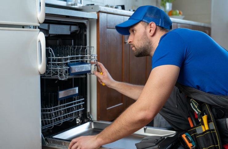 . A man in a blue shirt and hat works on a dishwasher, showcasing appliance repair services available in Sacramento, CA.