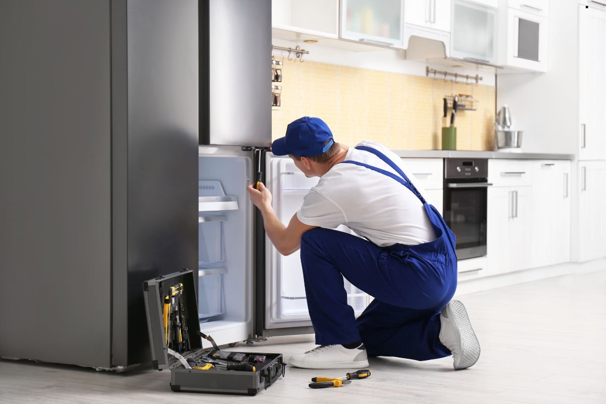 A technician in blue overalls crouches to repair a refrigerator in a modern kitchen, surrounded by tools and appliances. kitchen setting.