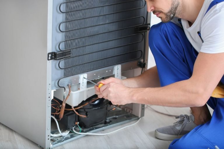 A technician in blue overalls repairs the back of a refrigerator, using a screwdriver near the coils and internal components.
