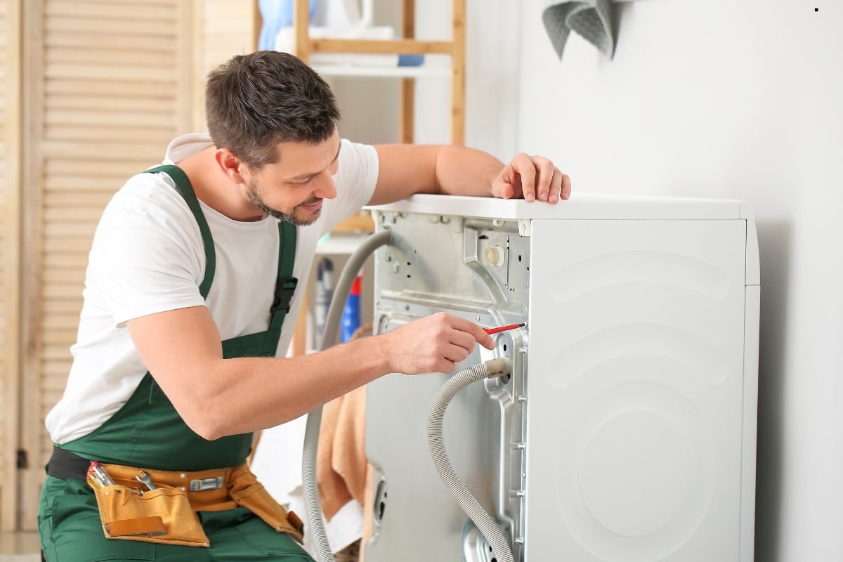 A technician in green overalls inspects the back of a washing machine, using a tool for maintenance in a bright laundry room.