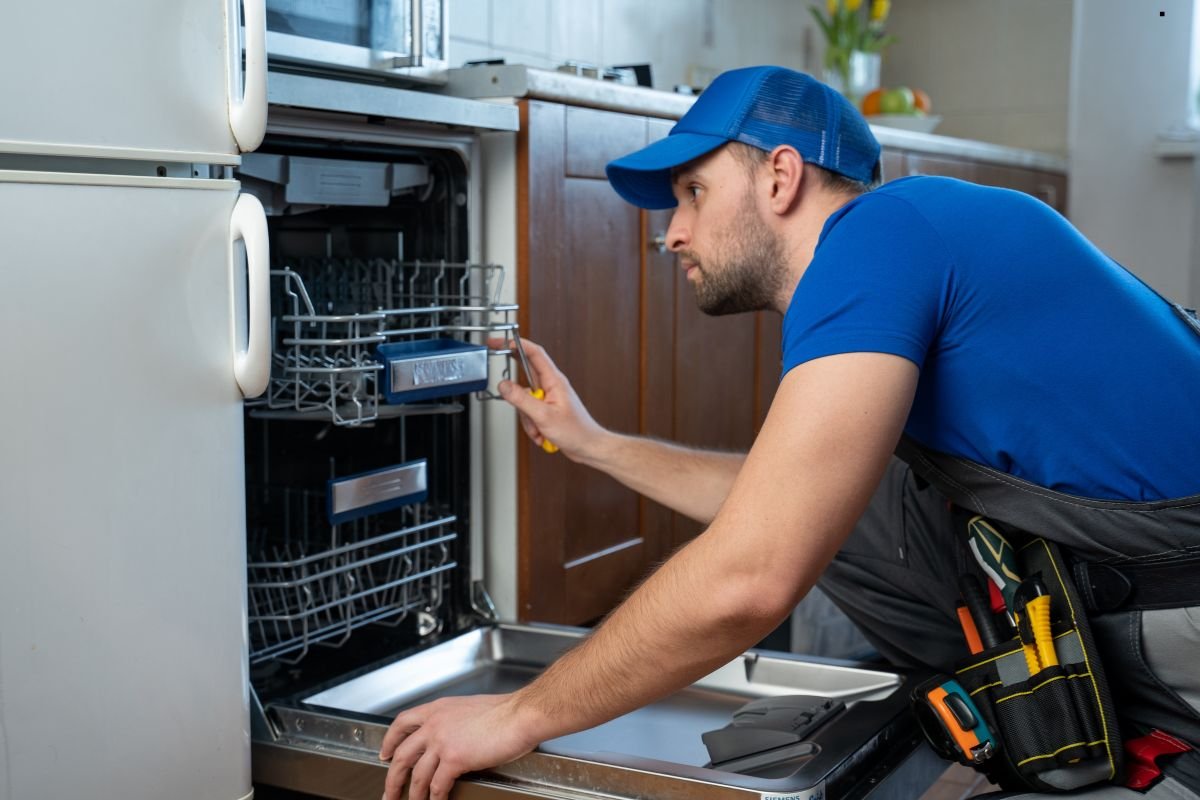 A technician in a blue shirt uses a tool to work on a dishwasher in a modern kitchen with wooden cabinets and a fridge.