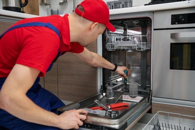 A technician in red shirt and blue overalls repairs a dishwasher, using tools and adjusting components inside the appliance.